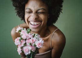 A cheerful woman with a radiant smile holding a pink carnation bouquet against a green background.